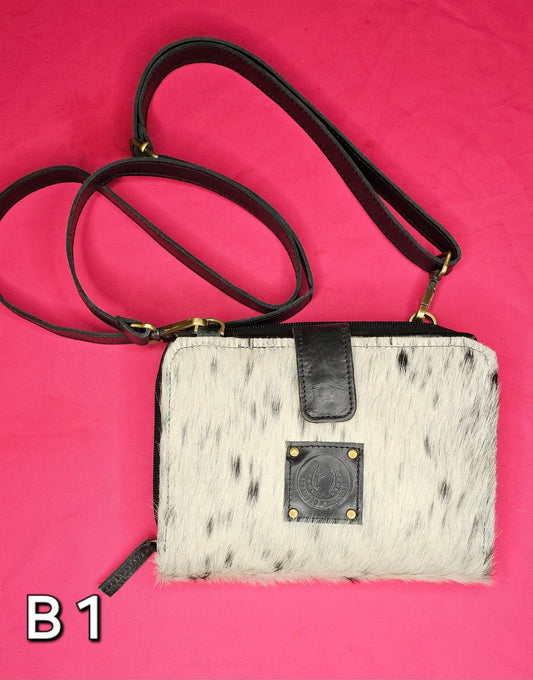 A black and white cowhide wallet with a shoulder strap and antique brass hardware, featuring the Branded Cowgirl logo.