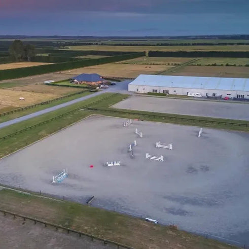 Aerial view of an equestrian arena with jumps and a building in the background.